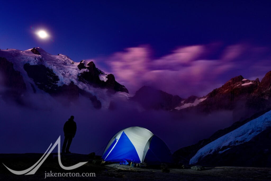 A lone tent perched at Turner’s Bivvy on the slopes of Mount Madeline, New Zealand. In the distance, a full moon rises over Mount Tutoko. 