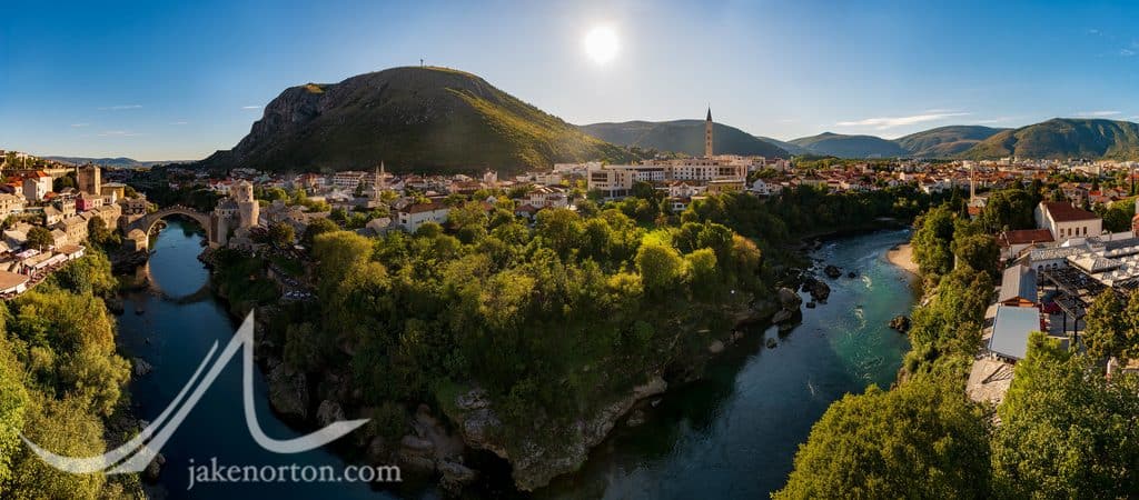 Mostar from above: Stari Most at left, the Neretva bending through town, the Millennium Cross atop Hum Hill, and the bell tower of the Franciscan church rising on the right.